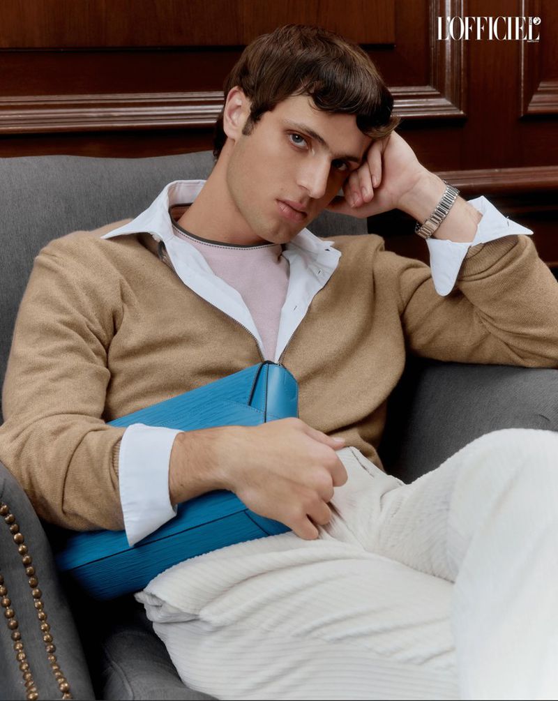Portrait of a brunette male model reclining on a grey chair, wearing a tan sweater, and cream pants, holding a teal bag.