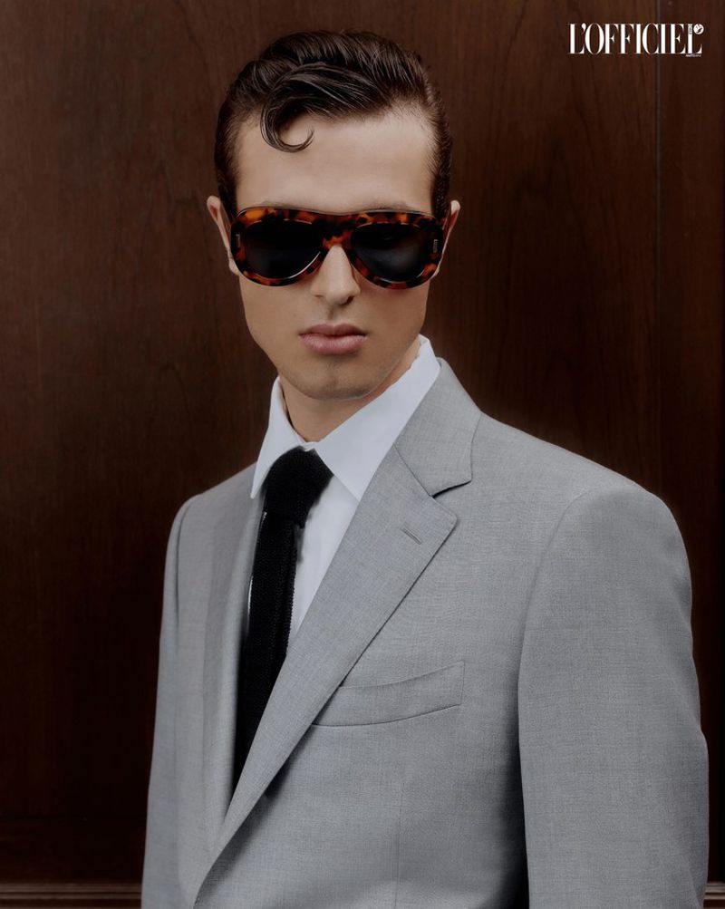 Man in a grey suit and black tie wearing tortoiseshell sunglasses stands against a dark wood background in a portrait orientation.