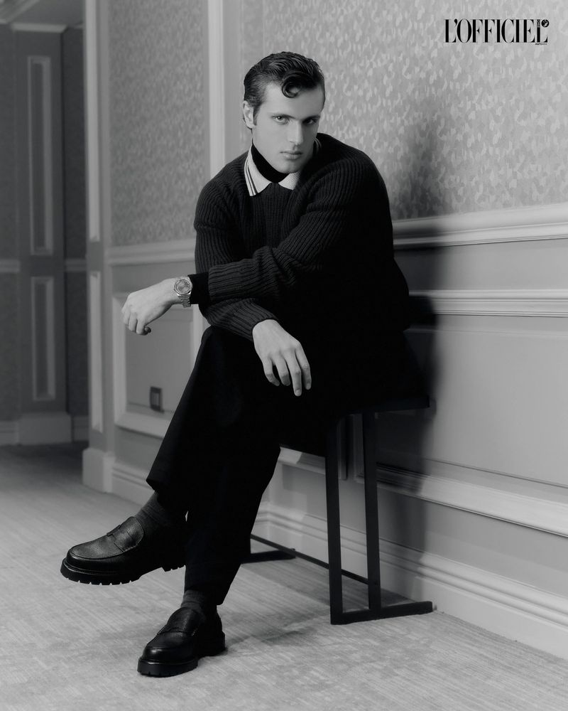 Black-and-white portrait of a handsome man sitting on a stool. He wears a dark sweater and pants with black loafers in an indoor setting.