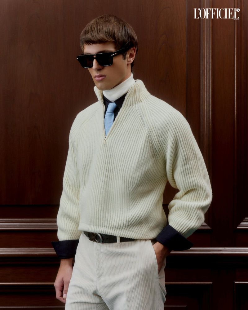 A stylish adult male poses indoors wearing a ribbed knit sweater, sunglasses, and light pants. Dark wood provides a rich backdrop.
