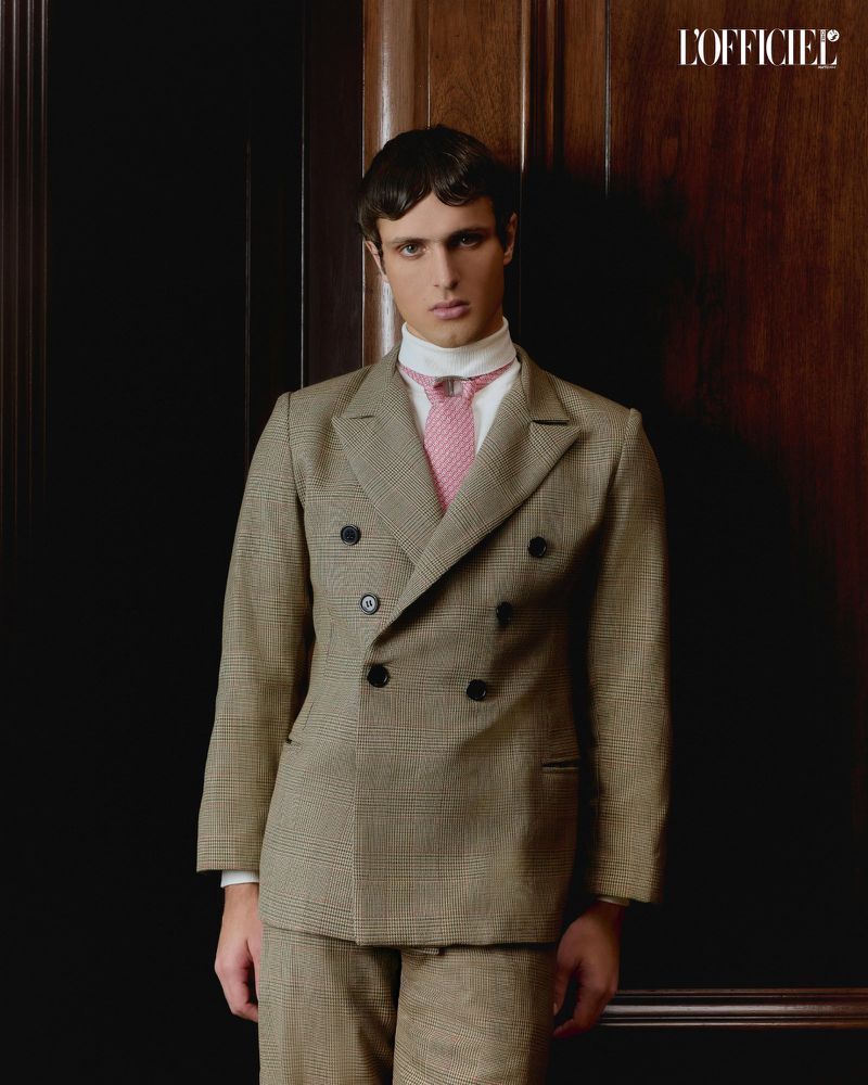 A well-dressed man in a double-breasted suit, collared shirt and tie, standing indoors in front of a dark wood paneled wall.