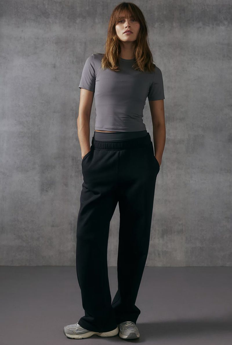 Studio shot of a woman posing in a grey t-shirt and black sweatpants, standing against a textured grey background.