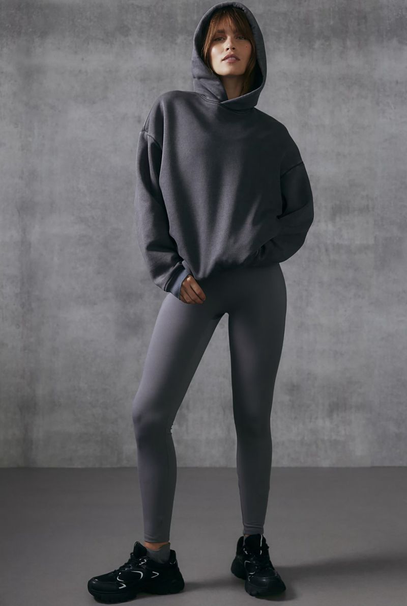 Woman in grey hooded sweatshirt and leggings with black sneakers stands against grey textured backdrop in a fashion shot.