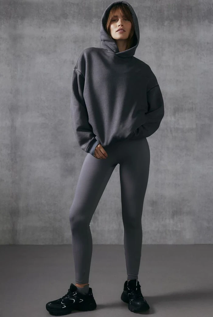 Model wearing a dark grey hoodie and matching leggings with black sneakers against a concrete textured wall.
