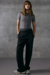 Model standing against a textured grey studio backdrop wearing a grey short-sleeve top and black sweatpants.