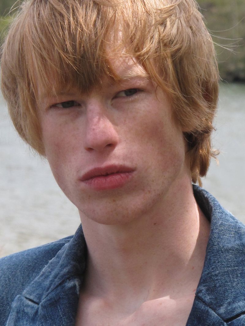 Close-up portrait of a fair-skinned young man with ginger hair and freckles wearing a blue denim jacket, outdoors by a water surface.