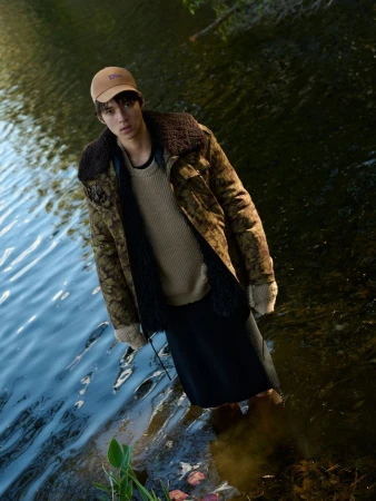 Man wearing a brown patterned jacket, beige sweater, and baseball cap standing in rippling outdoor water.