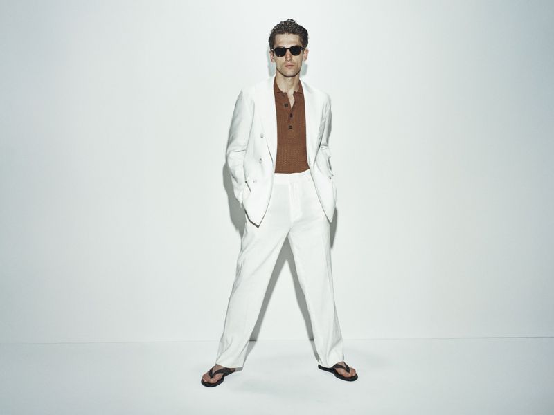 Man in white suit and sunglasses poses in photo studio. He is wearing a brown shirt and black sandals against a white backdrop.