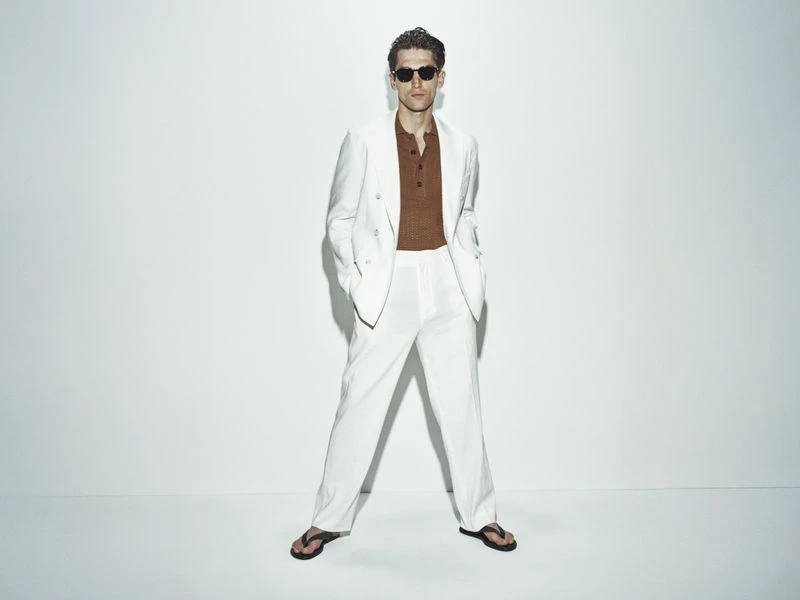 Man in white suit and sunglasses poses in photo studio. He is wearing a brown shirt and black sandals against a white backdrop.
