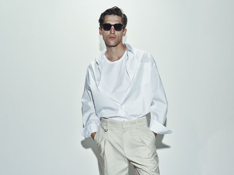 Portrait of a stylish adult man in a white shirt and beige pants with sunglasses posing against a neutral background in a studio setting.