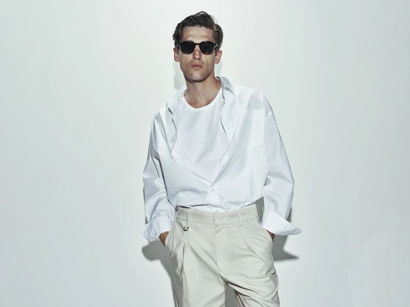 Portrait of a stylish adult man in a white shirt and beige pants with sunglasses posing against a neutral background in a studio setting.