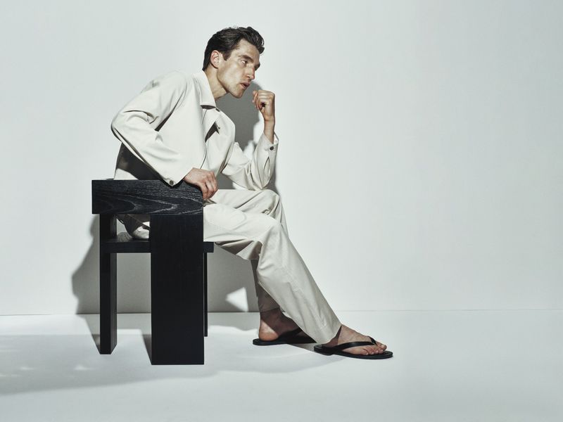 Adult man in a tan outfit sitting on a black chair in a light photo studio setting, hand near his face, looking off-camera.
