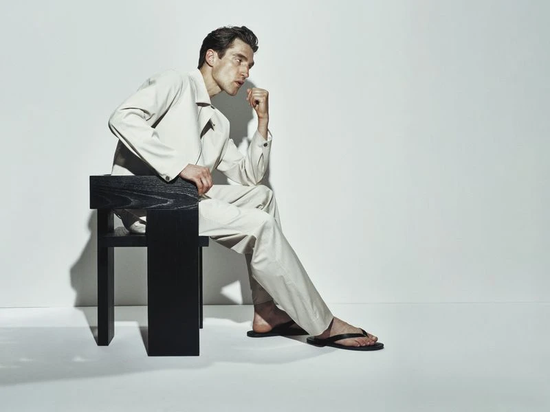 Adult man in a tan outfit sitting on a black chair in a light photo studio setting, hand near his face, looking off-camera.