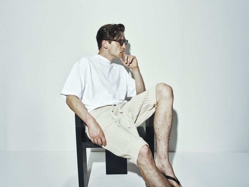 A man wearing sunglasses and a white t-shirt sits on a chair in a studio. He has short dark hair and wears a pair of beige shorts.