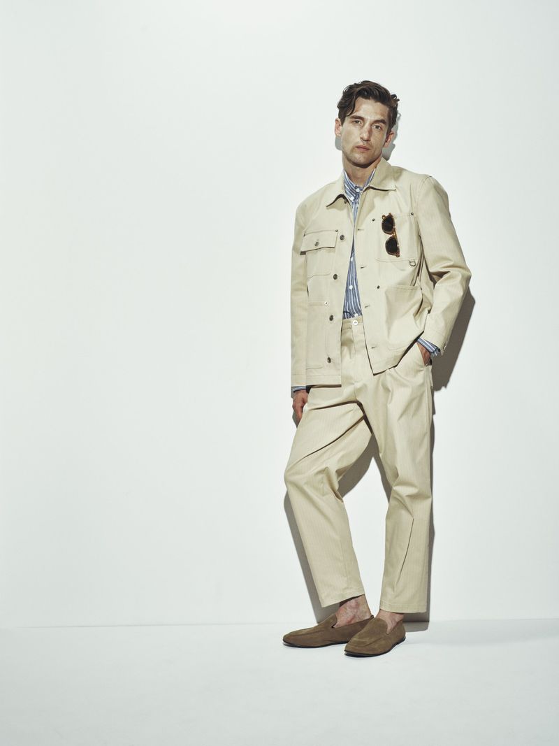 A stylish adult male poses against a bright white backdrop, wearing a tan jacket and pants, with sunglasses and brown loafers.