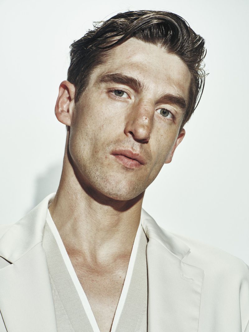 Portrait of a young adult male wearing a blazer and neutral-colored shirt, gazing into the camera against a white background in a studio.