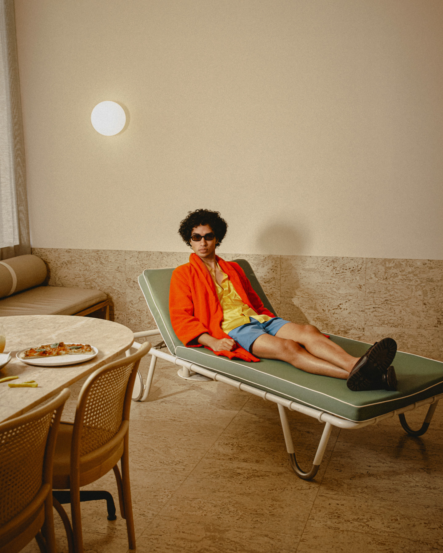 A man wearing an orange robe and sunglasses reclines on a green sun lounger indoors beside a table with pizza.