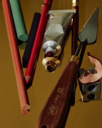 Art supplies including an open paint tube, palette knife, colored pencils, and sharpener arranged on a reflective gold surface.