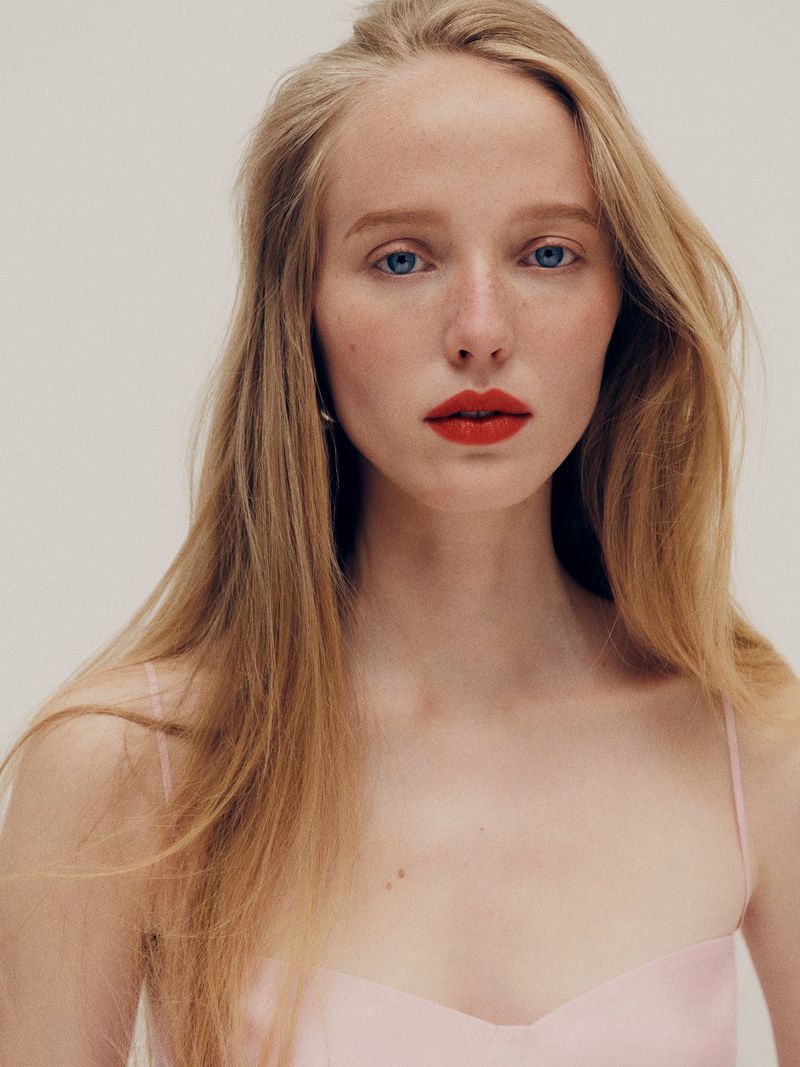 Close-up portrait of model Maelle Swaelens with long blonde hair and bold red lipstick wearing a pale pink spaghetti-strap top.