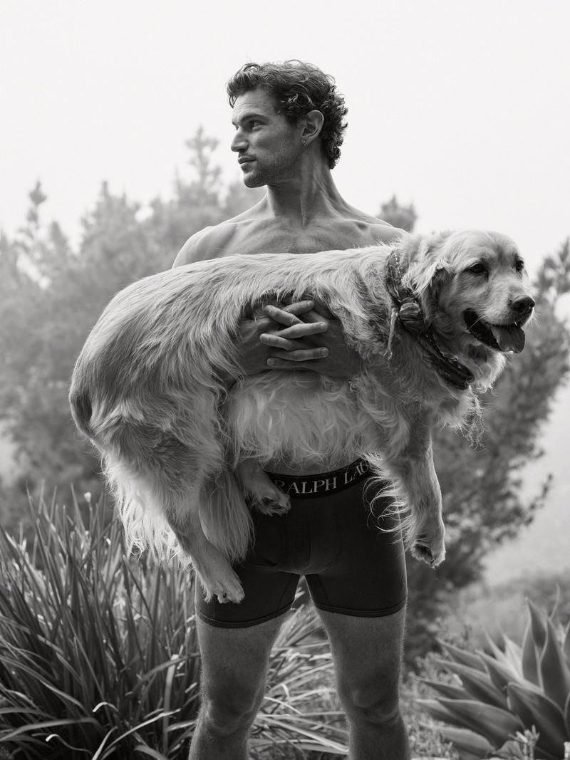 A shirtless man standing outdoors holding a large golden retriever in his arms while wearing black Polo Ralph Lauren boxer briefs.