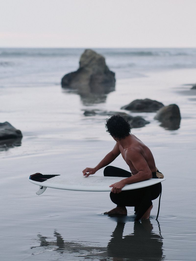 A man in black wetsuit pants squatting on a wet sandy beach holding a white surfboard, photographed from behind.