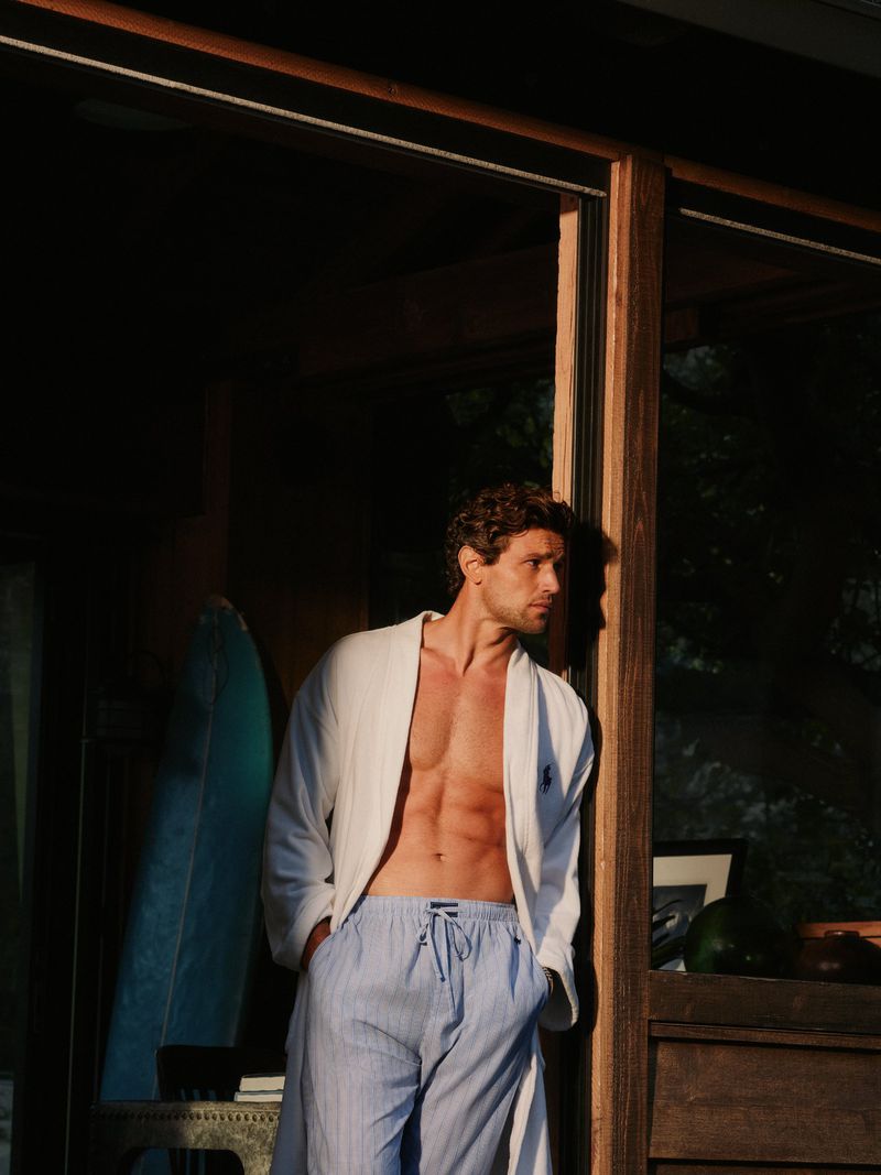 Model Giacomo Cavalli leaning in a doorway wearing an open white bathrobe and light blue lounge pants in warm sunlight.