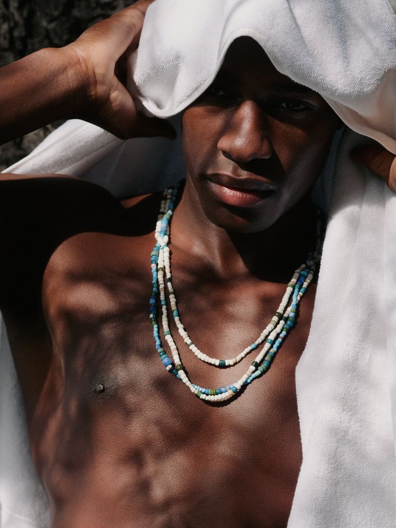 Shirtless Black male model holding a white towel over his head and wearing layered beaded necklaces under direct sunlight.