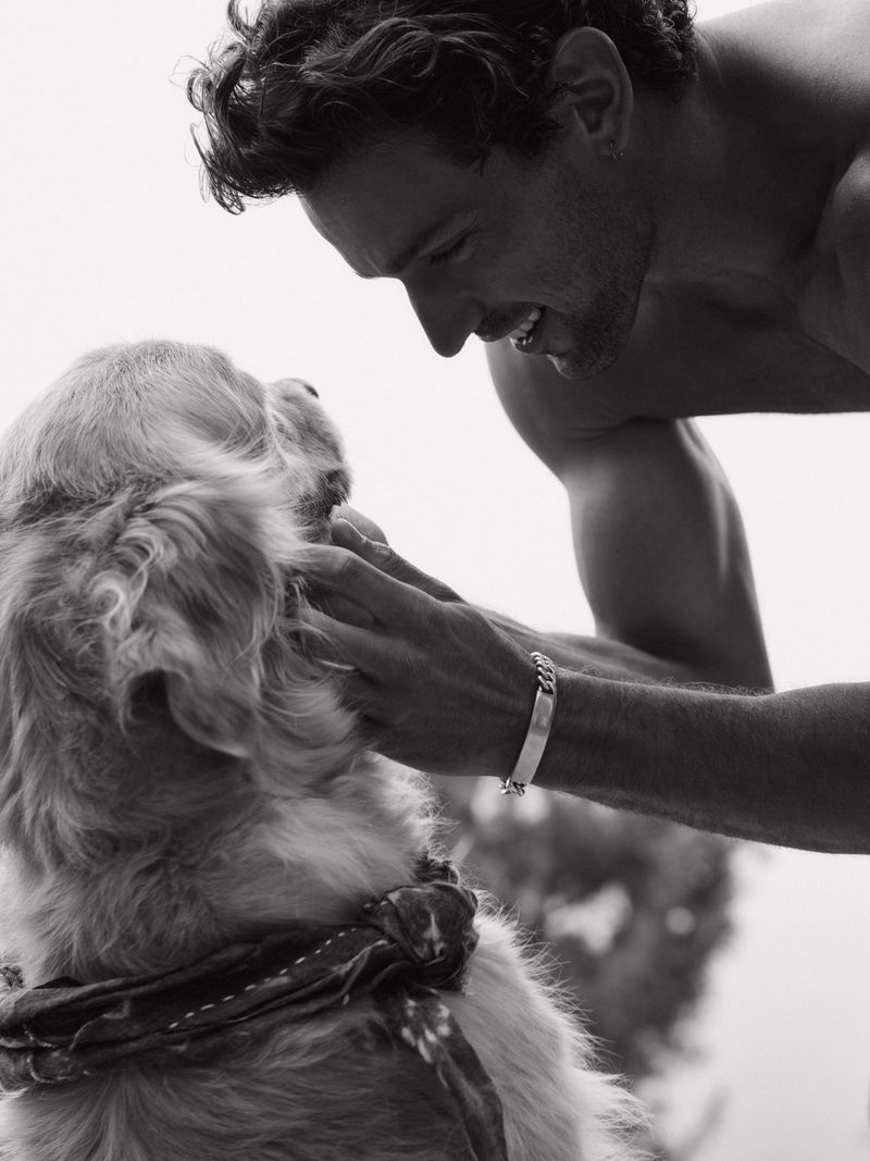 Black and white photo of a shirtless man smiling warmly while petting a golden retriever wearing a neckerchief outdoors.