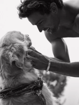Black and white photo of a shirtless man smiling warmly while petting a golden retriever wearing a neckerchief outdoors.