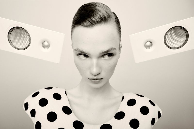 High-key studio portrait of a woman in a black and white polka dot top flanked by two white audio speakers against a light backdrop.