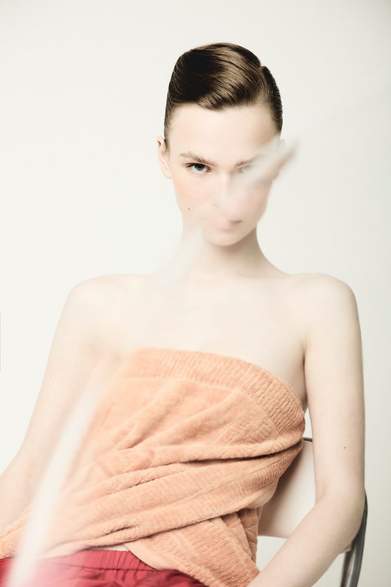 Model with slicked hair wrapped in a peach terrycloth towel and red shorts against a plain light studio background.