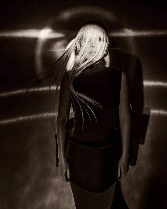Monochrome portrait of a model with windblown blonde hair wearing a black sleeveless mini dress against projected circular lighting.