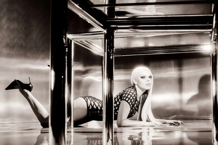 Model with platinum blonde hair lying beneath a chrome table wearing a black lattice bodysuit and stiletto heels.