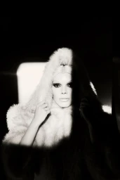 Black and white portrait of a woman wearing a plush hooded fur coat with high-contrast chiaroscuro lighting cutting across her face.