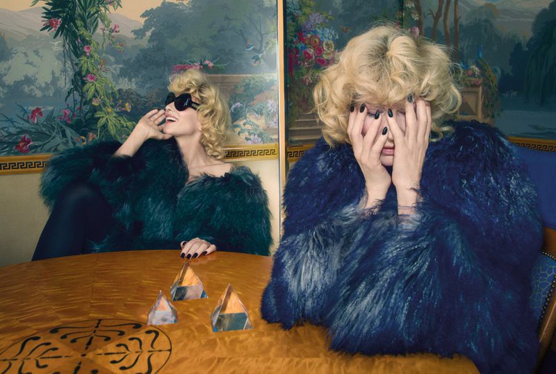 Blonde woman wearing a blue faux-fur coat sitting at a polished wooden table in a room with ornate painted screen panels