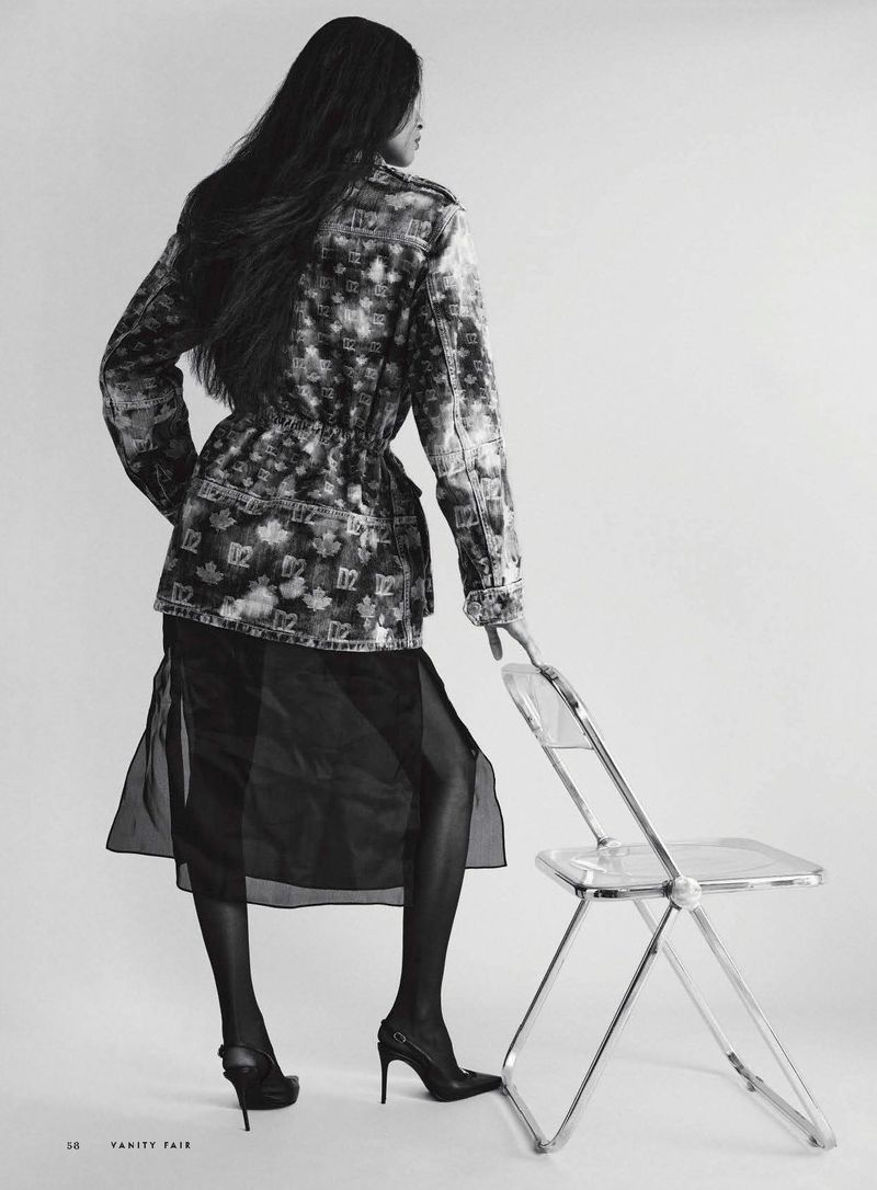 Model standing facing away in a patterned jacket, sheer black skirt, and pointed slingback stiletto heels beside a clear folding chair.