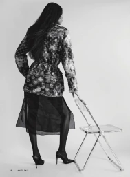 Model standing facing away in a patterned jacket, sheer black skirt, and pointed slingback stiletto heels beside a clear folding chair.