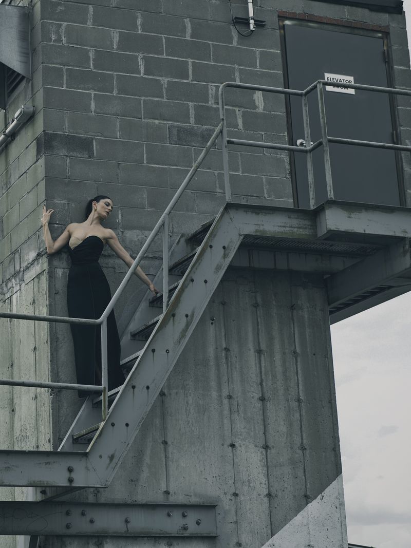 Monica Barbaro in a strapless black jumpsuit poses against a brutalist grey concrete structure with an external staircase.
