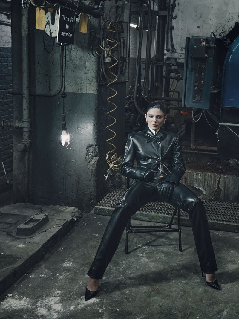 Monica Barbaro seated on a chair in a dark industrial boiler room wearing a black leather tailored jacket, trousers, and leather gloves.