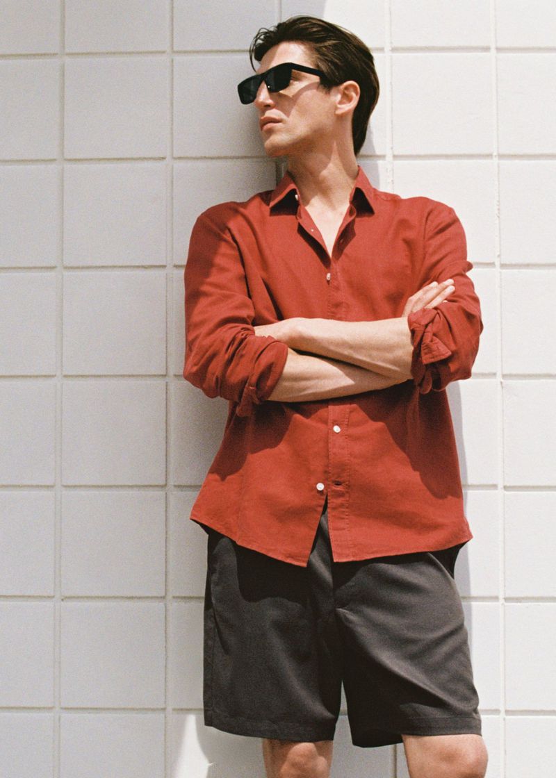 A stylish adult man wearing sunglasses, a red collared shirt, and gray shorts stands against a tiled wall in natural outdoor light.