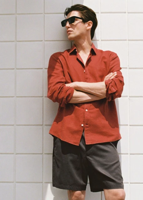A stylish adult man wearing sunglasses, a red collared shirt, and gray shorts stands against a tiled wall in natural outdoor light.