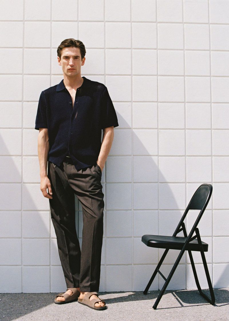 A full-length portrait of a man wearing a navy polo shirt and brown trousers stands next to a black folding chair against a white tiled wall.