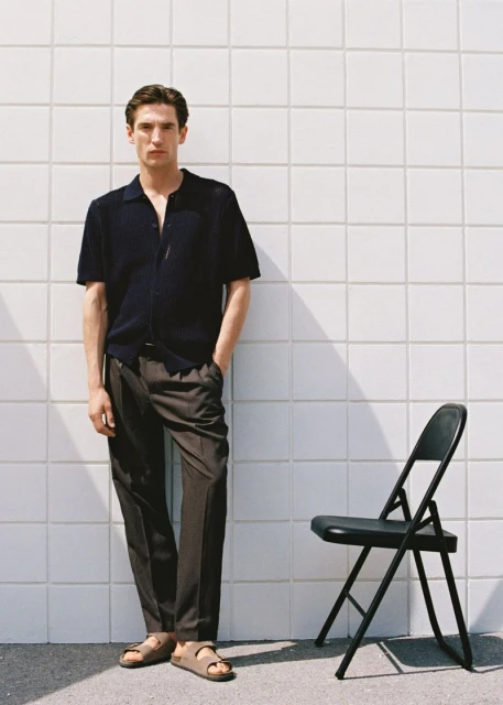 A full-length portrait of a man wearing a navy polo shirt and brown trousers stands next to a black folding chair against a white tiled wall.
