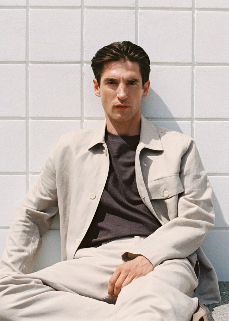 Portrait of a young adult man in a light beige linen suit sitting cross-legged against a white tiled wall in natural light.