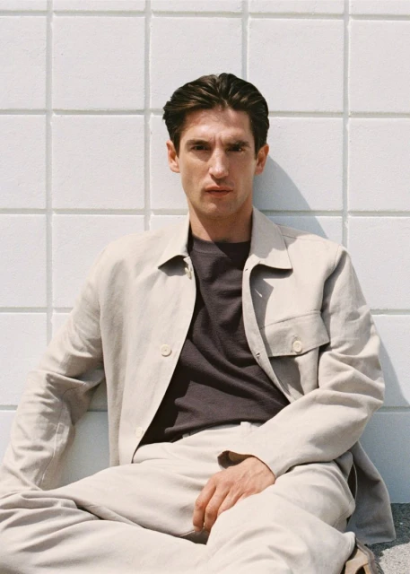 Portrait of a young adult man in a light beige linen suit sitting cross-legged against a white tiled wall in natural light.
