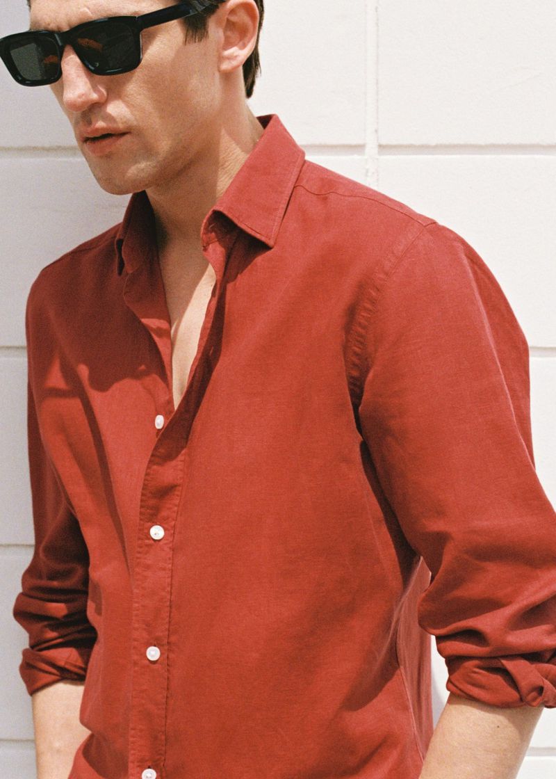 Head and shoulders shot of adult male wearing a red button-down shirt and dark sunglasses, standing against a white wall.