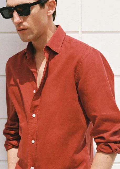 Head and shoulders shot of adult male wearing a red button-down shirt and dark sunglasses, standing against a white wall.