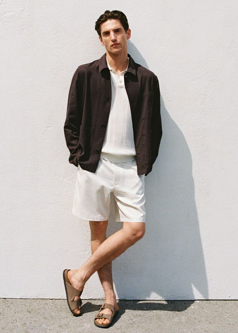 Male fashion model poses in neutral tones, wearing a brown jacket, white polo, shorts, and sandals against a light-colored wall.