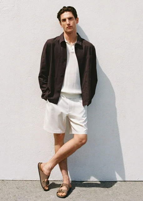 Male fashion model poses in neutral tones, wearing a brown jacket, white polo, shorts, and sandals against a light-colored wall.