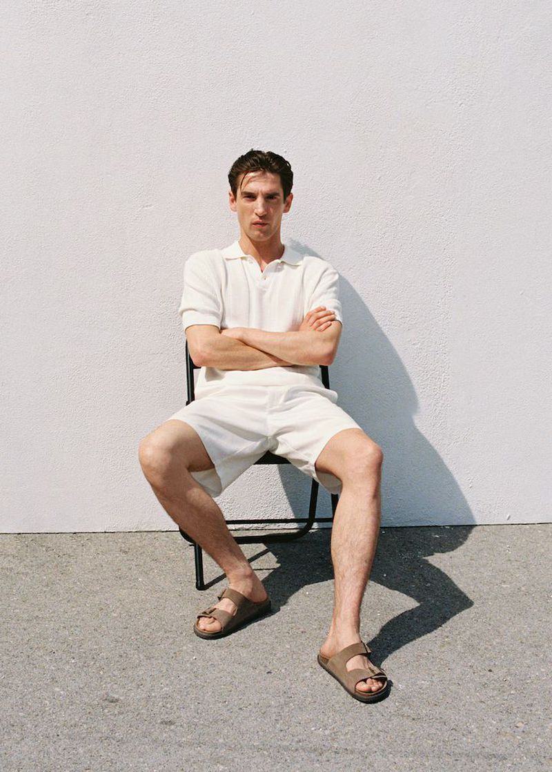 Adult male in off-white polo and shorts sitting on a chair outside, wearing sandals, with arms crossed against a white wall.